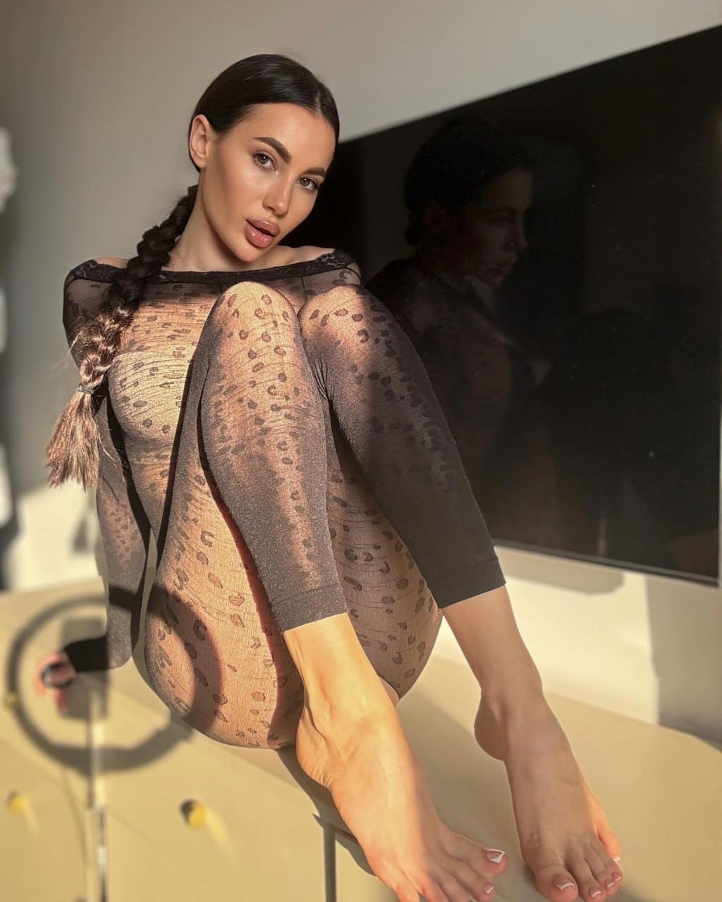 A woman with a long braid sits on a stool in a sheer leopard-print bodysuit, feet on the base, against a dark screen.