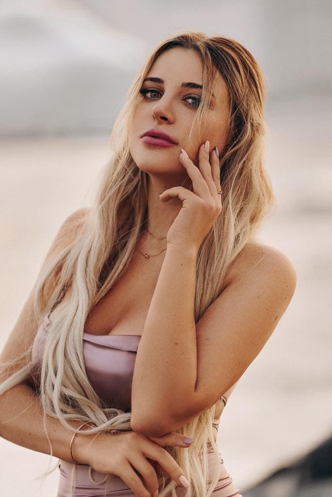 Milana with long wavy blonde hair stands outdoors in a light pink sleeveless top, hand raised to her cheek, against a soft-focus natural background.