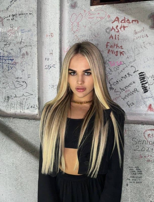 Nita stands in a black cropped top and jacket with a gold choker, facing the camera against a graffiti-covered wall.