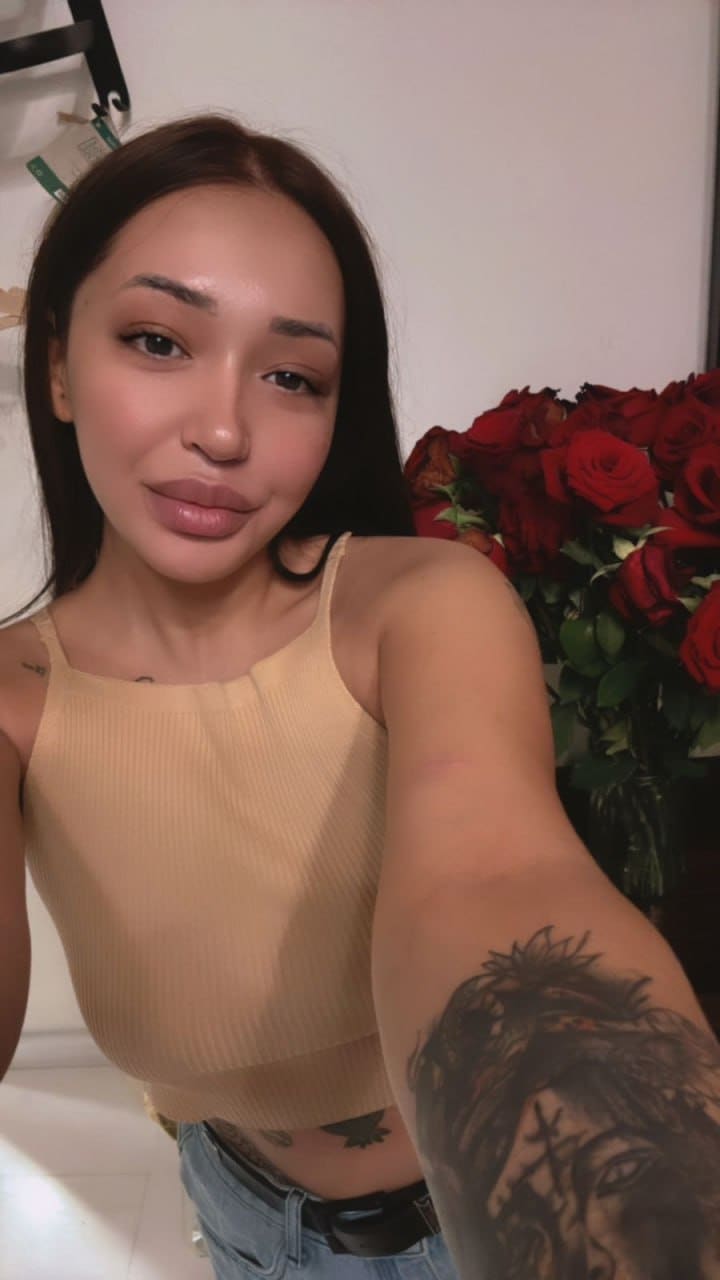 Nella in a beige ribbed tank top and light blue jeans, showing a forearm tattoo, with red roses behind her against a white wall.