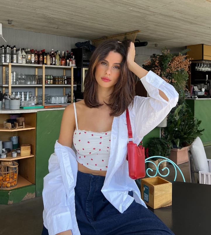 Seated at a cafe table before shelves of bottles, she wears a white polka-dot camisole, jeans, and an open shirt with a red bag.