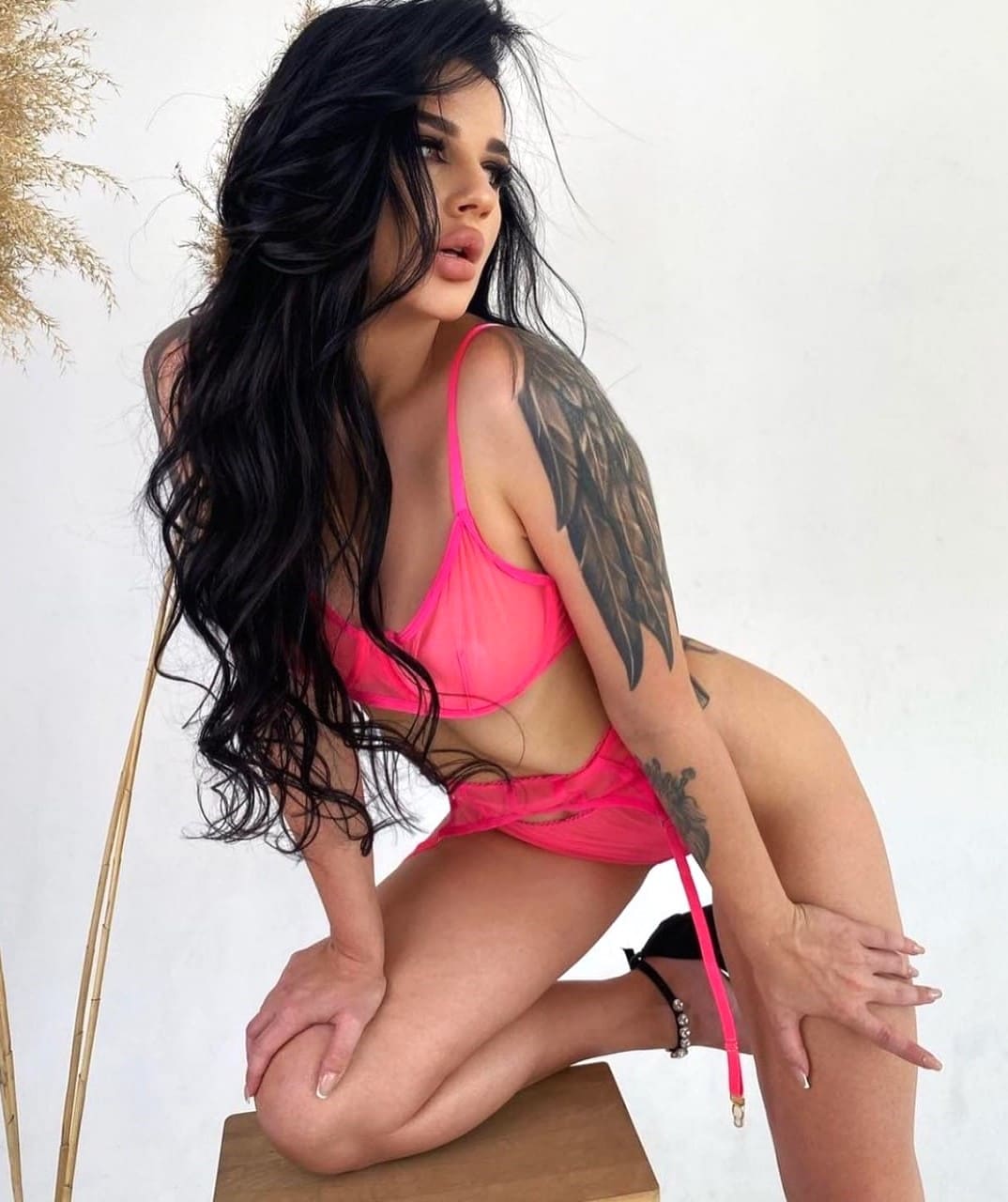 A woman kneels on a wooden stool in a pink bikini against a white wall, with pampas grass to the left and a wing tattoo visible.