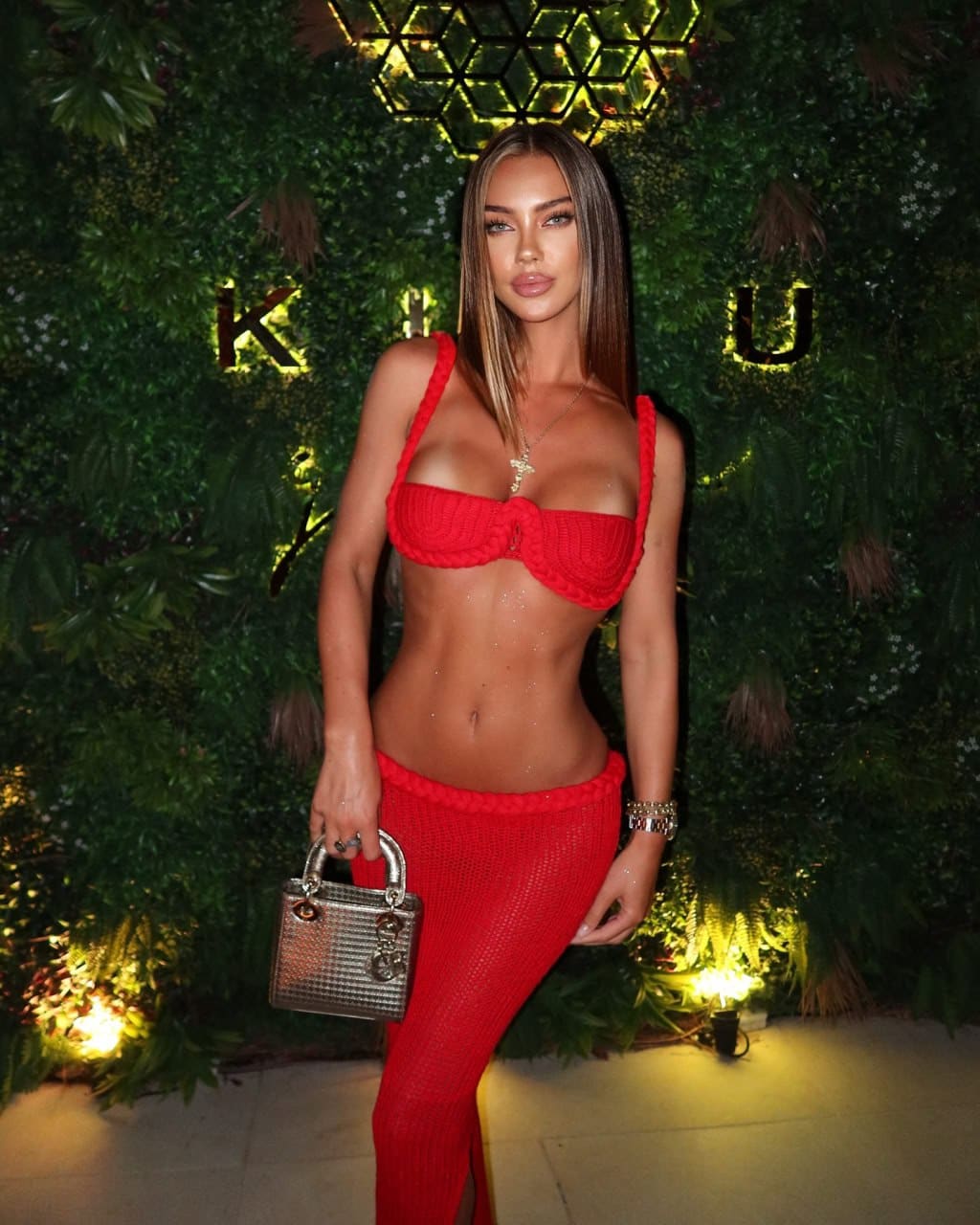 Nancy stands in a red cropped tube top and high-waisted pants, holding a metallic bag before a green foliage wall with lit letters.