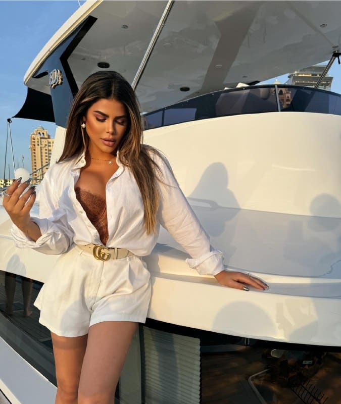 On a boat deck, a woman in an unbuttoned white shirt, brown lace top, and white shorts rests a hand on the edge.