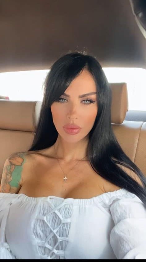 A woman with a shoulder tattoo sits in a car back seat wearing a white lace-up top and cross necklace, facing forward.