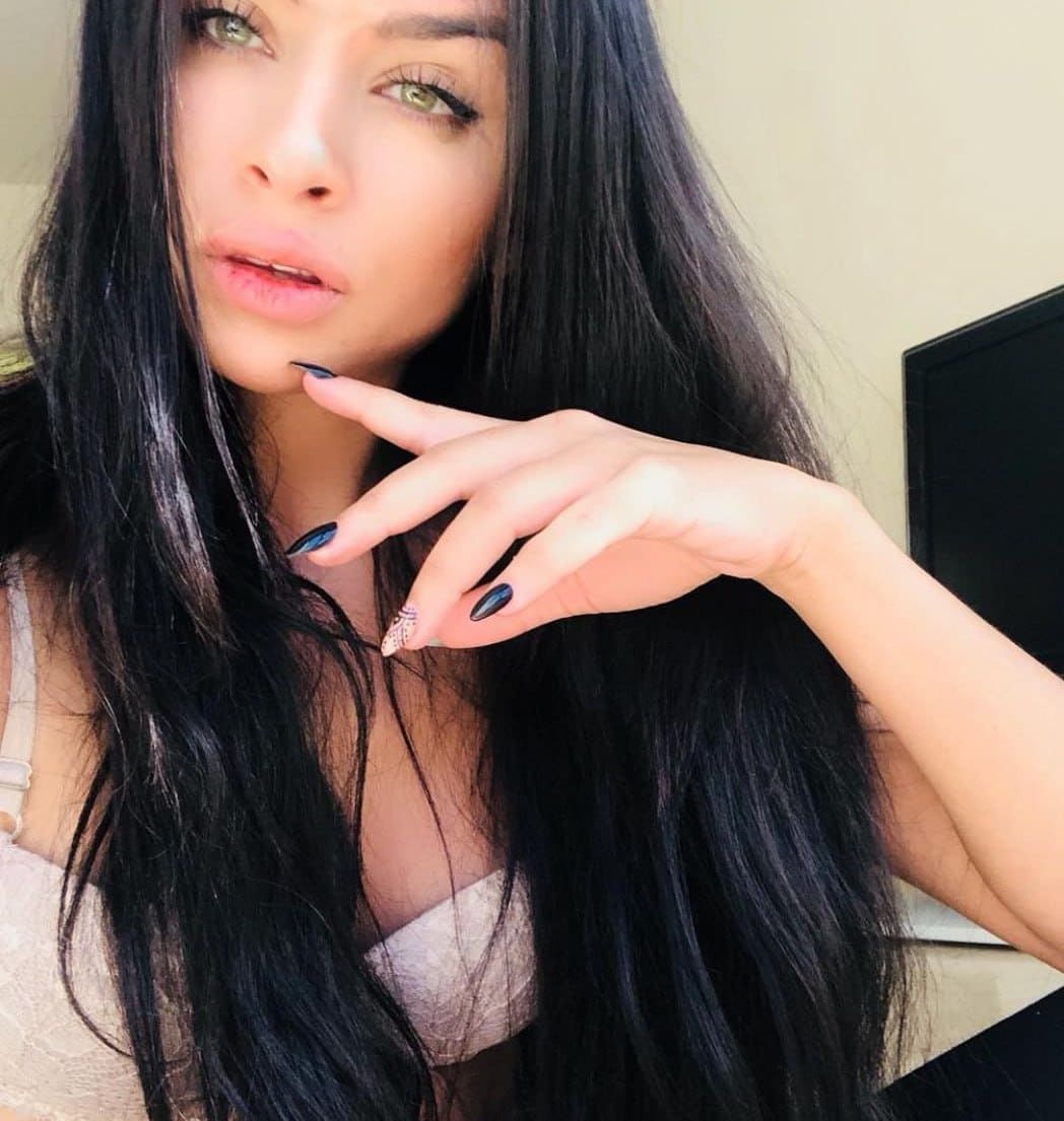 A woman with dark nail polish rests her hand near her chin, wearing a light lace top indoors against a plain wall and dark-framed object.