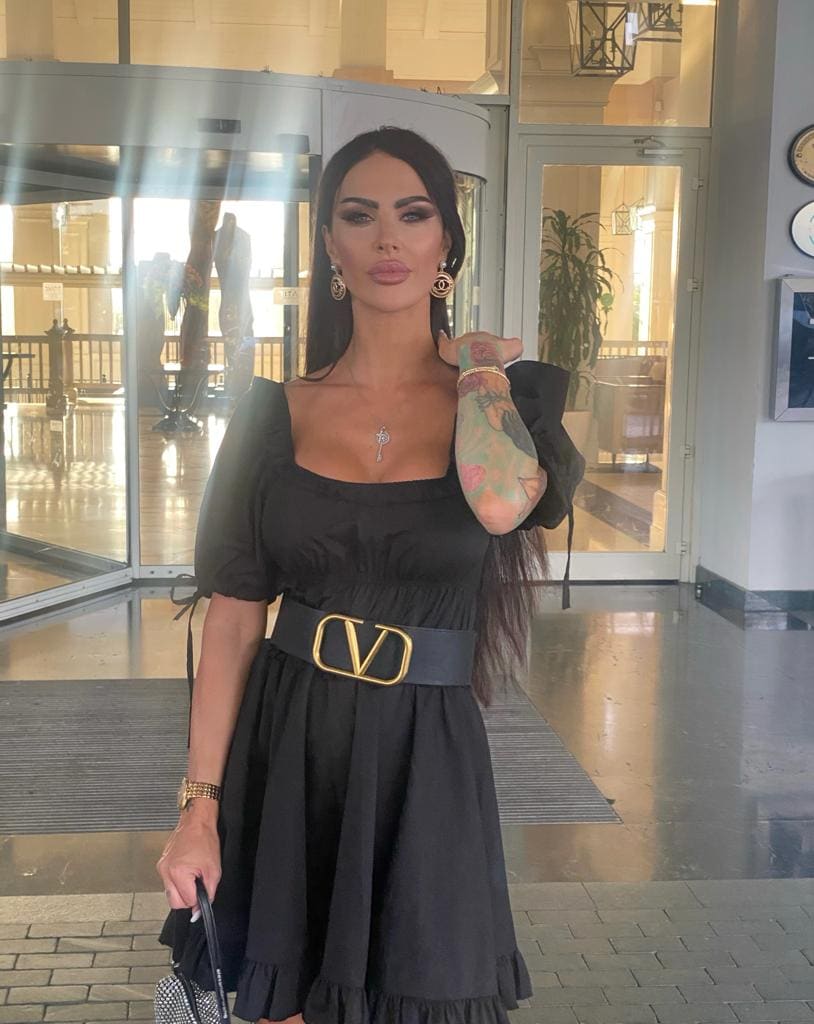 Abelle stands in a hotel lobby wearing a black dress with a gold-buckled belt, holding a bag and raising her tattooed arm.