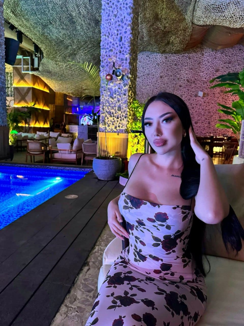 Violet sits on a chair by a blue-lit pool, wearing a sleeveless floral dress with her hand on her head.