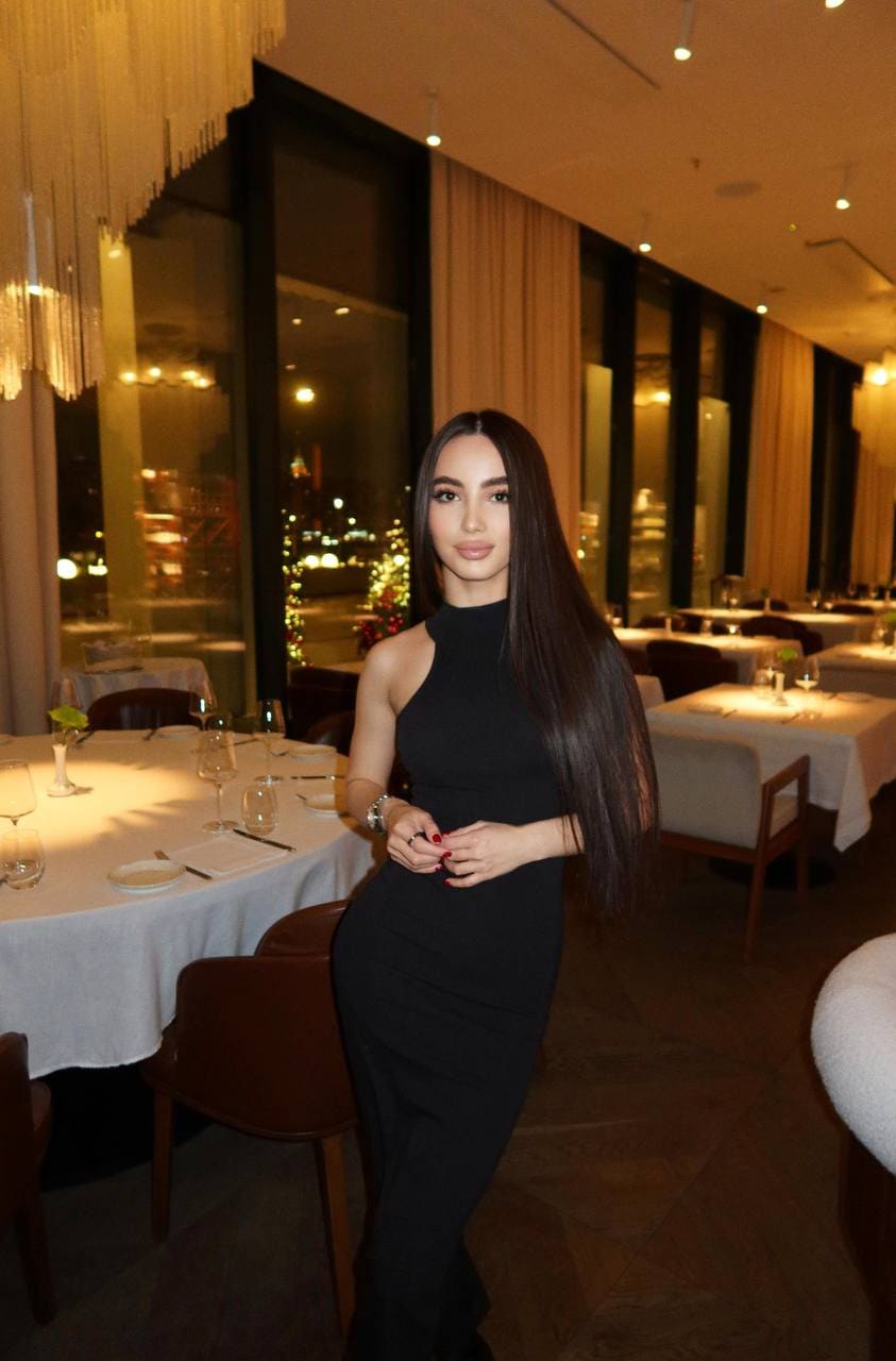 A woman with long dark hair stands in a restaurant wearing a sleeveless black high-neck dress, hands clasped near a set white table.