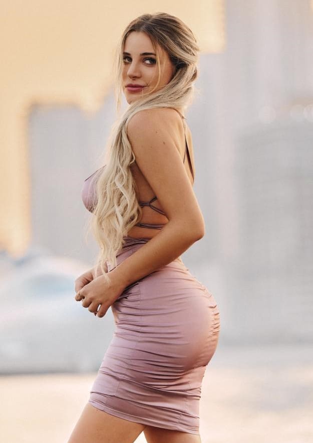 Profile view of a woman in a tight, backless pink mini dress, hand on hip, standing before a blurred urban background.