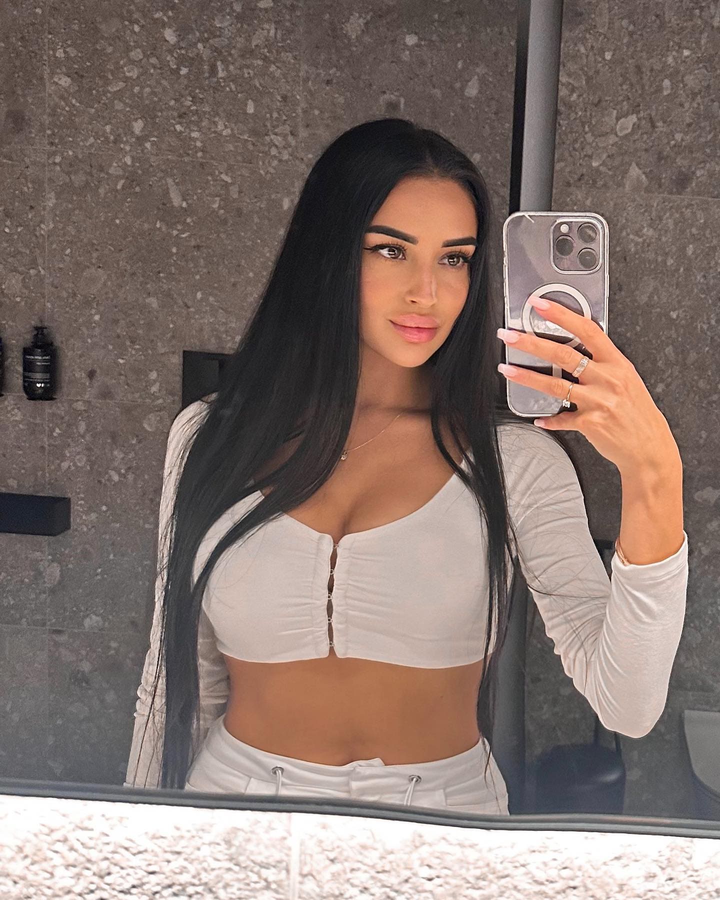 A mirror selfie shows a woman in a white cropped button-up top and drawstring pants, holding a smartphone in a bathroom.