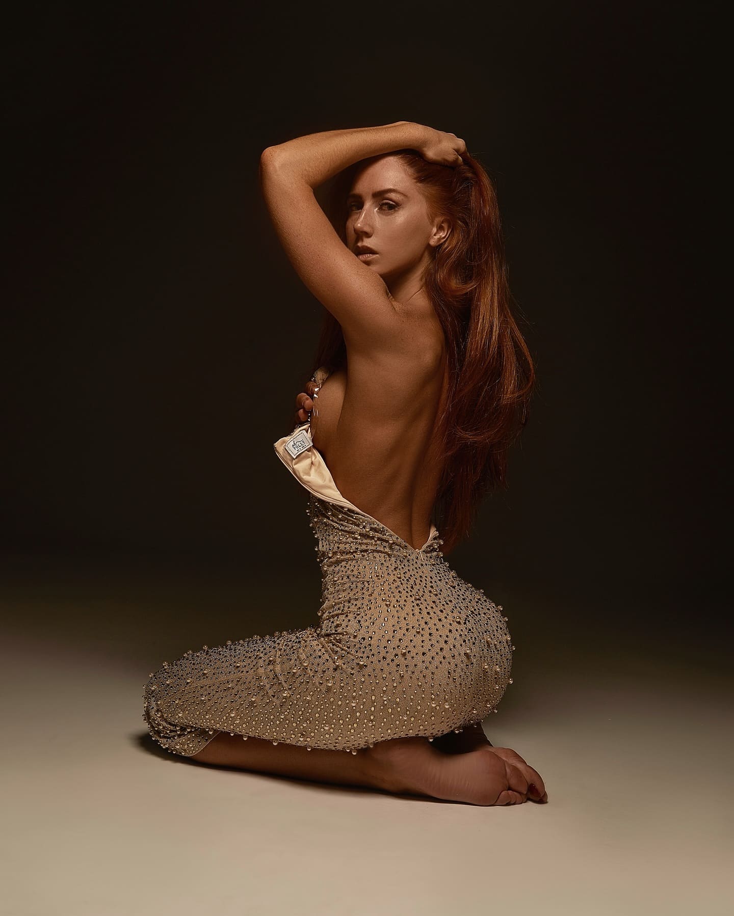 A kneeling woman wears a backless beaded dress, holding the top edge with one hand and resting the other on her head against a dark background.