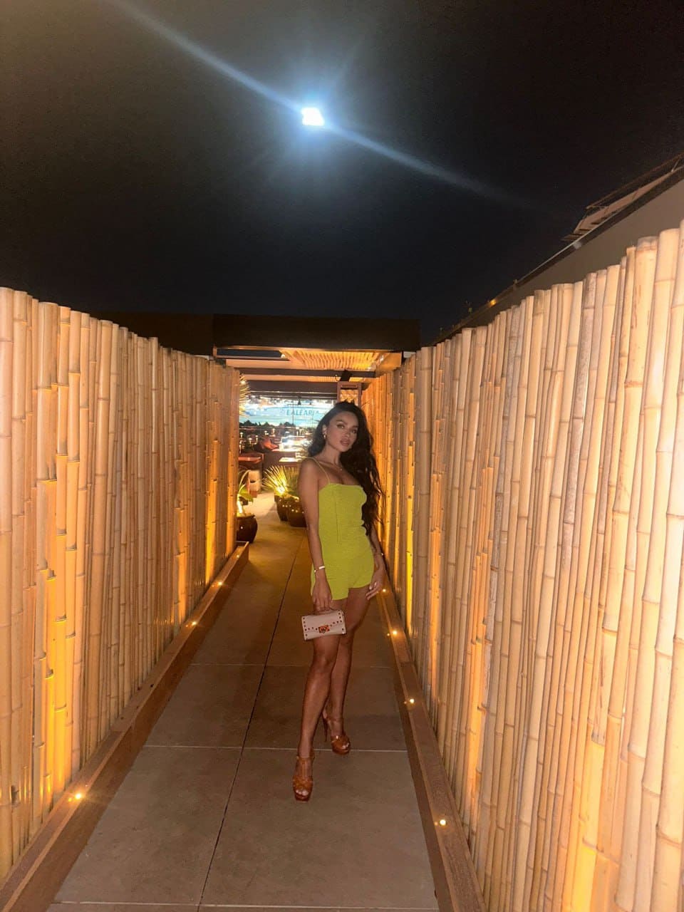 A woman in a lime-green strapless dress and brown heels stands in a bamboo corridor, holding a small white clutch in her hand.