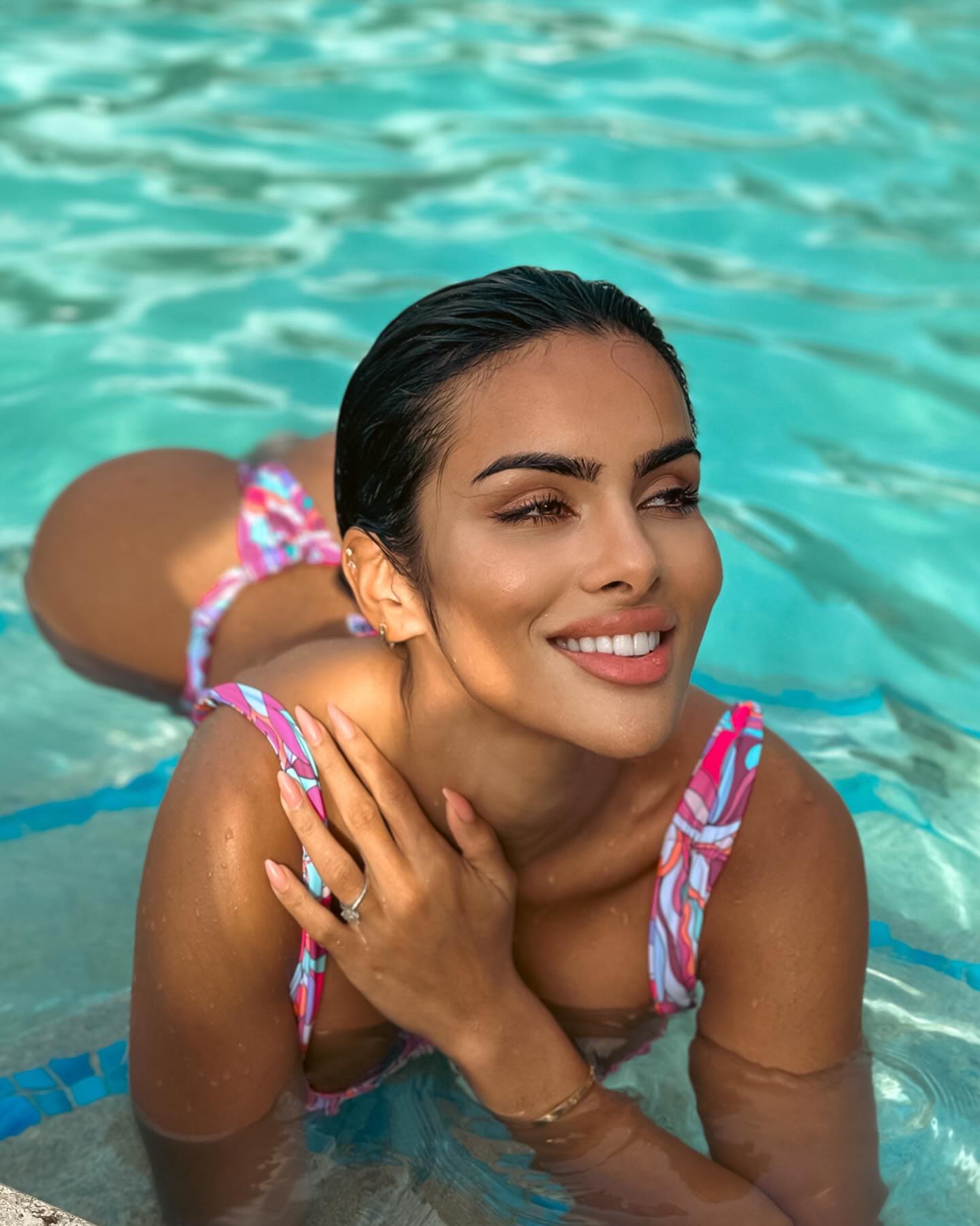 Izzy lies in a turquoise pool wearing a pink and blue patterned two-piece swimsuit, one hand on her shoulder, smiling at the camera.