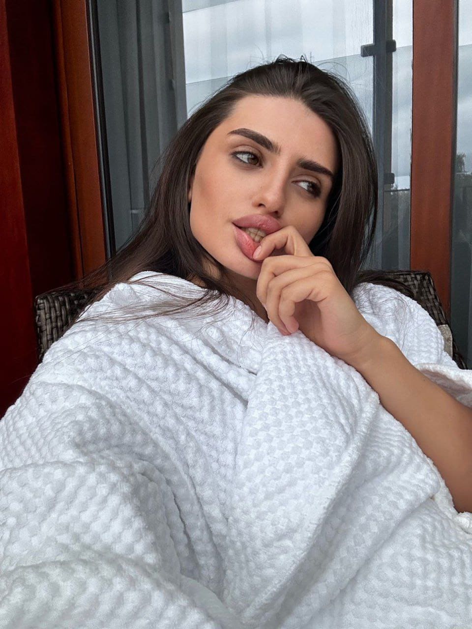 Aliana sits in a woven chair wrapped in a white blanket, hand to mouth, looking aside near red curtains.