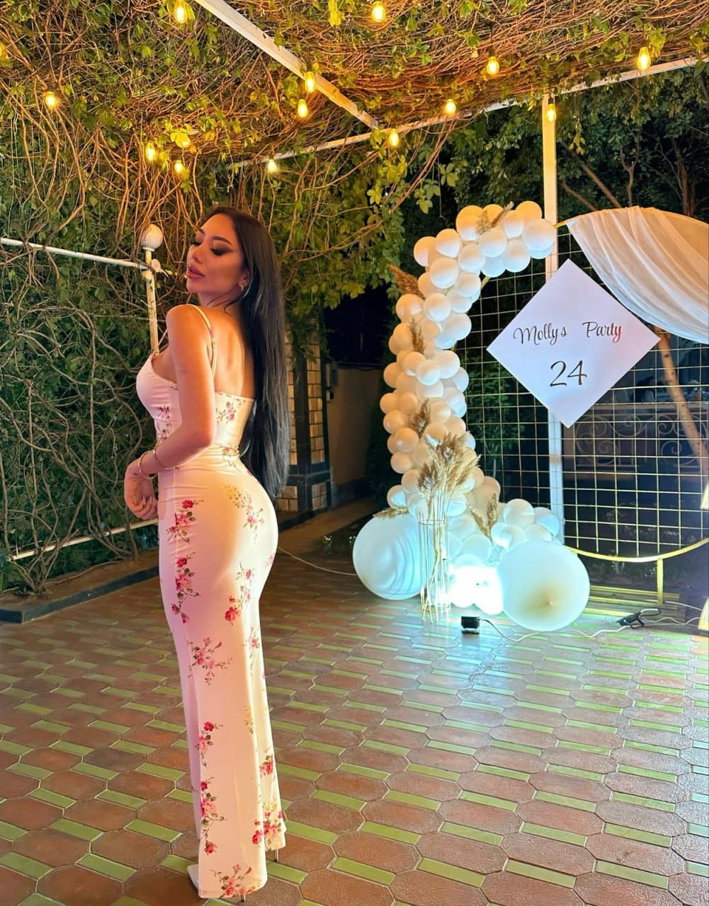 A woman in a long pink floral dress stands in profile on a patio near a white balloon arch and string lights.